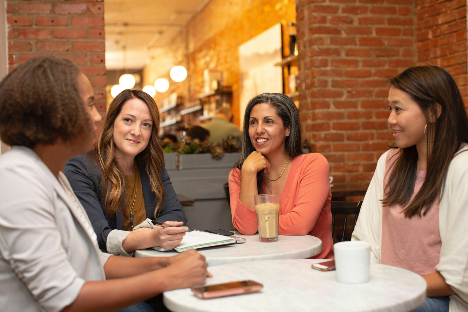 Stuents and a mentor sitting together at a table, engaged in a discussion. One student is holding a notebook and pen, while the mentor gestures with their hand, offering guidance. Both are smiling, indicating a positive and supportive interaction.